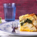Creamy spinach and cheese stuffed bread pudding with broccoli florets, served on a decorative plate with a fork, water glass, and mortar and pestle in the background for a rustic, comforting dish.