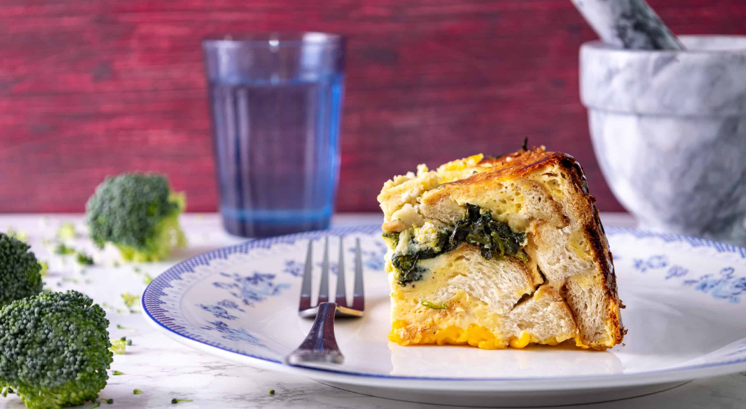 Creamy spinach and cheese stuffed bread pudding with broccoli florets, served on a decorative plate with a fork, water glass, and mortar and pestle in the background for a rustic, comforting dish.