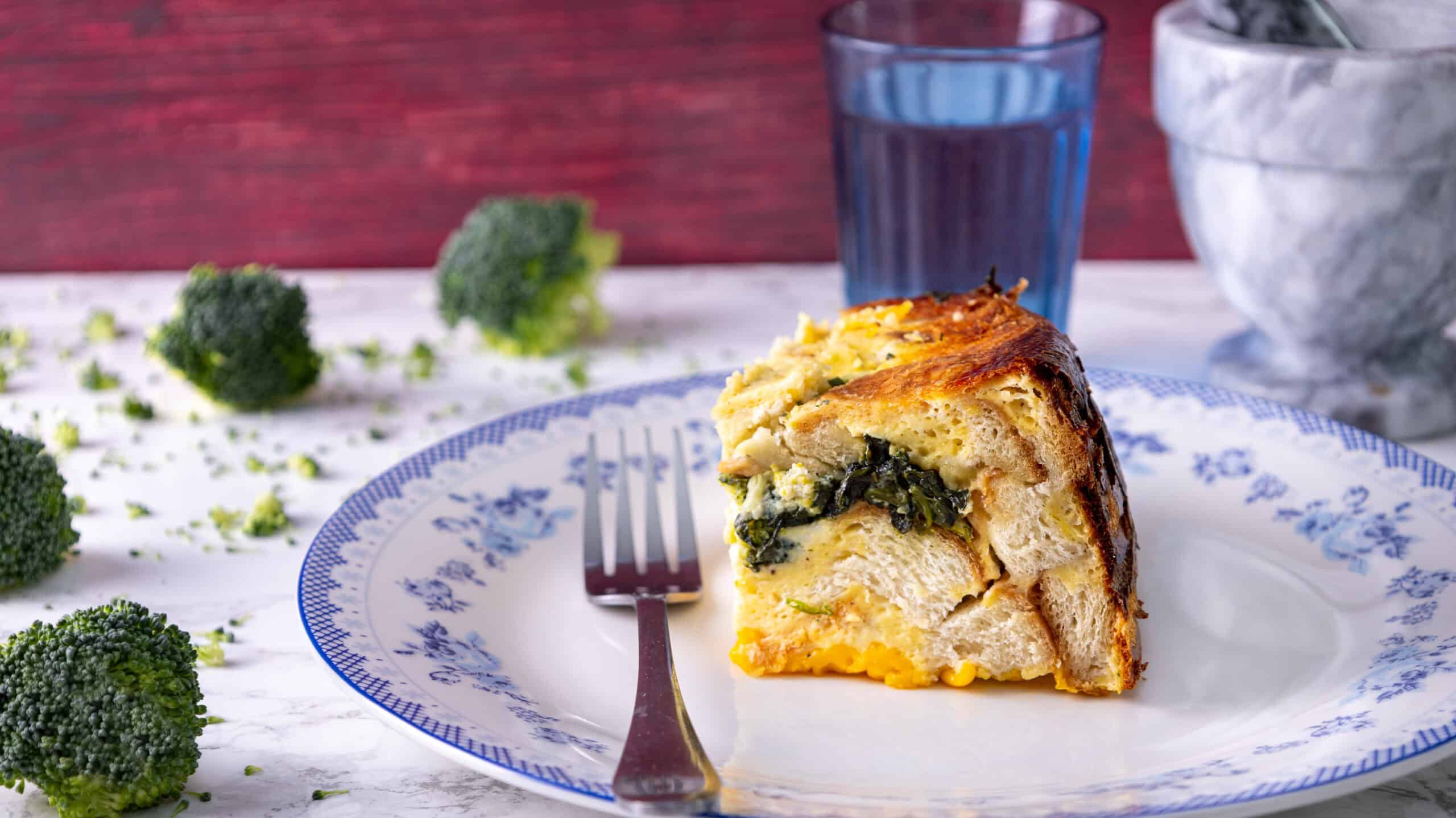 Broccoli and cheese stuffed chicken pot pie with golden crust on a white plate with blue floral design, served with a fork and a glass of water.