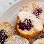 Homemade vanilla and berry muffins dusted with powdered sugar, served on a white plate with a decorative lace cloth. Perfect for breakfast or dessert, featuring fresh berries inside moist muffins.