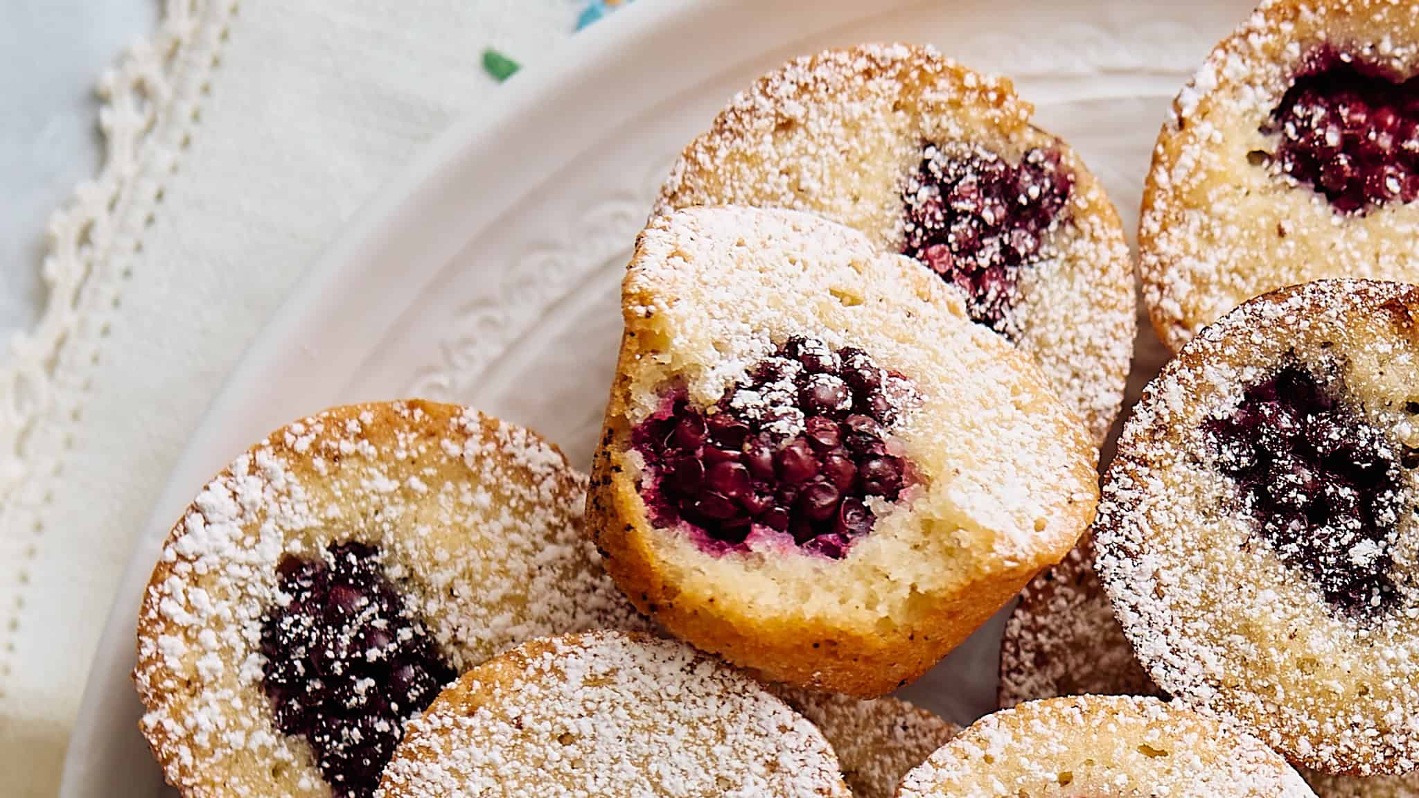 Homemade vanilla and berry muffins dusted with powdered sugar, served on a white plate with a decorative lace cloth. Perfect for breakfast or dessert, featuring fresh berries inside moist muffins.