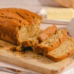 Moist homemade banana bread sliced on a wooden cutting board, with butter and cream cheese in the background, perfect for breakfast or snack.