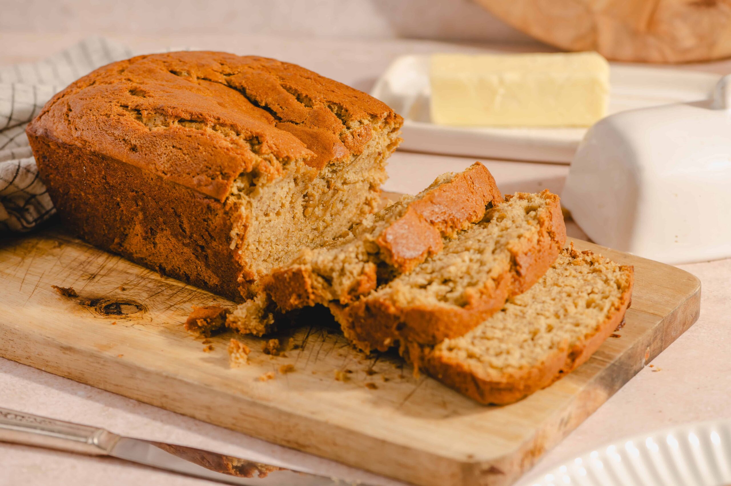 Moist homemade banana bread sliced on a wooden cutting board, with butter and cream cheese in the background, perfect for breakfast or snack.
