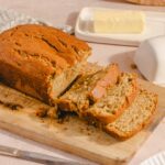 Moist banana bread loaf with slices on a wooden cutting board, butter and milk in the background, perfect for breakfast or snack.