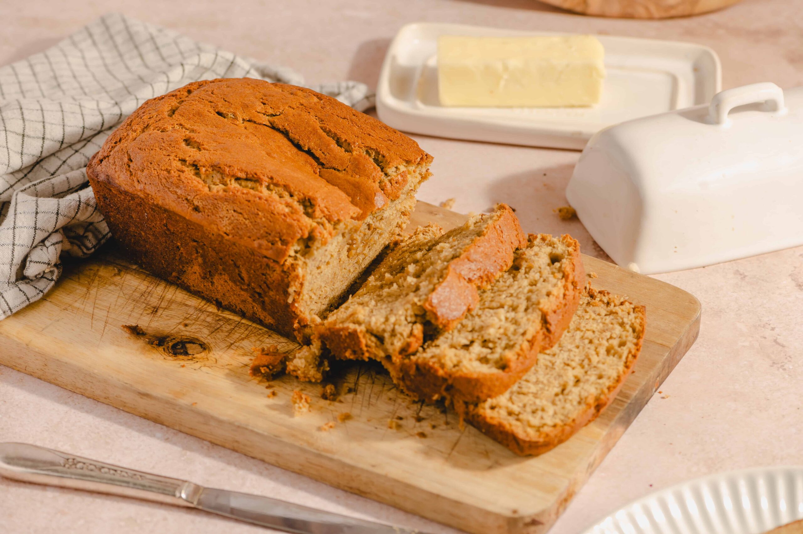 Moist banana bread loaf with slices on a wooden cutting board, butter and milk in the background, perfect for breakfast or snack.