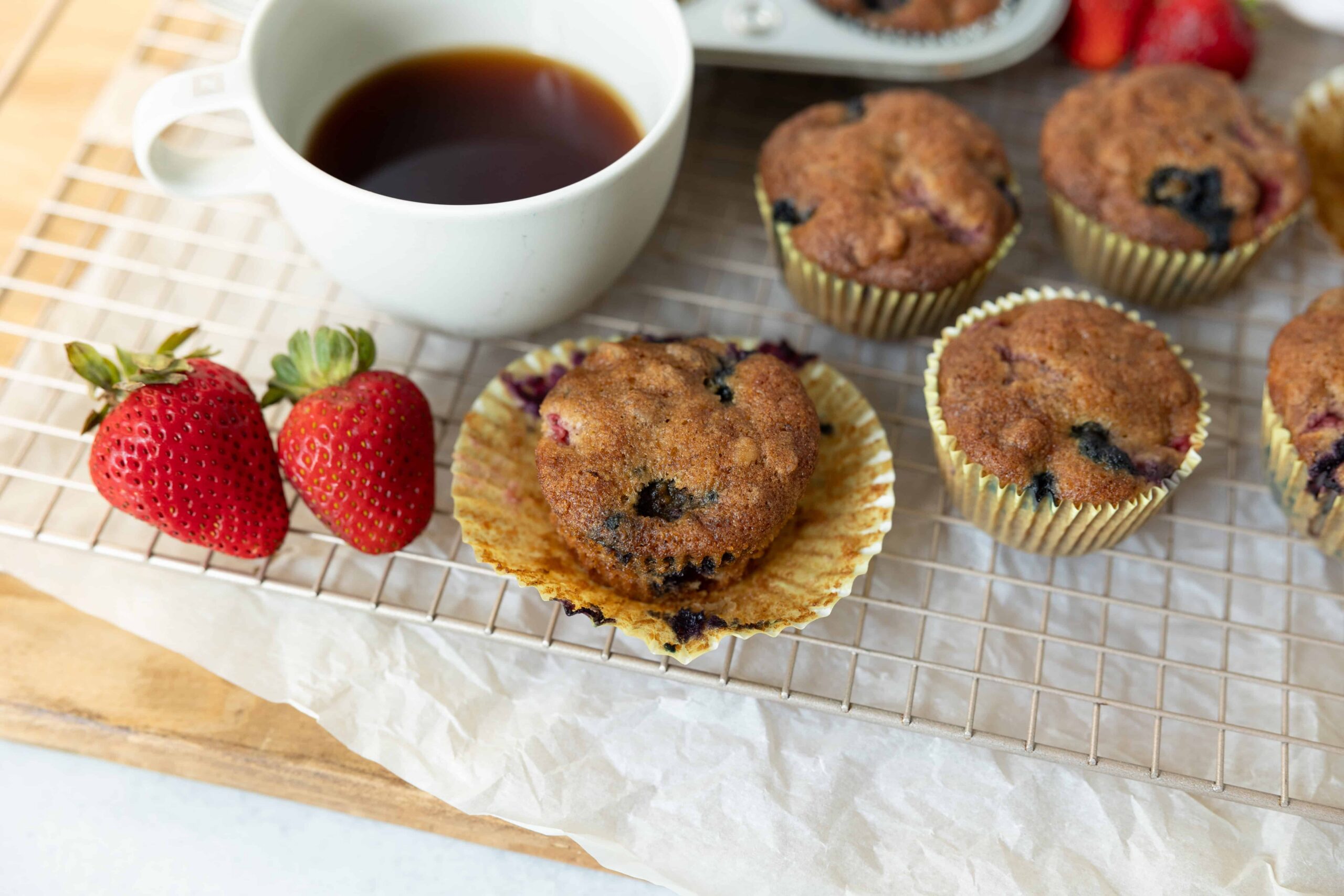 Fresh homemade blueberry muffins with strawberries and a cup of black coffee, served on a wire cooling rack for breakfast or snack. Perfect for berry lovers and baking enthusiasts.