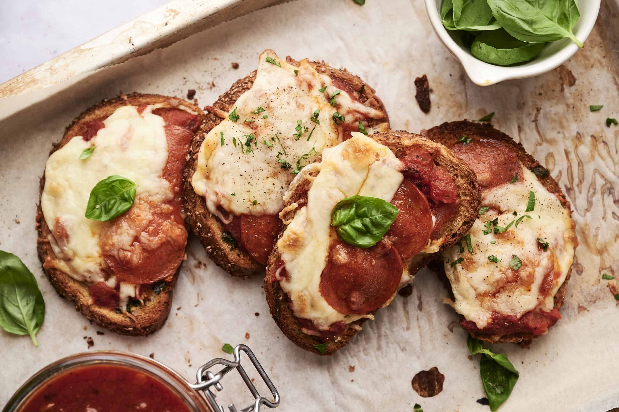 Oven-baked pepperoni and cheese mini pizzas on rustic bread slices with fresh basil garnish, served on parchment paper with a side of tomato sauce and a bowl of fresh spinach leaves, perfect for homemade pizza night.