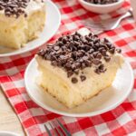 Creamy vanilla sponge cake topped with rich chocolate chips, served on a white plate with a red checkered tablecloth, perfect for dessert or teatime.
