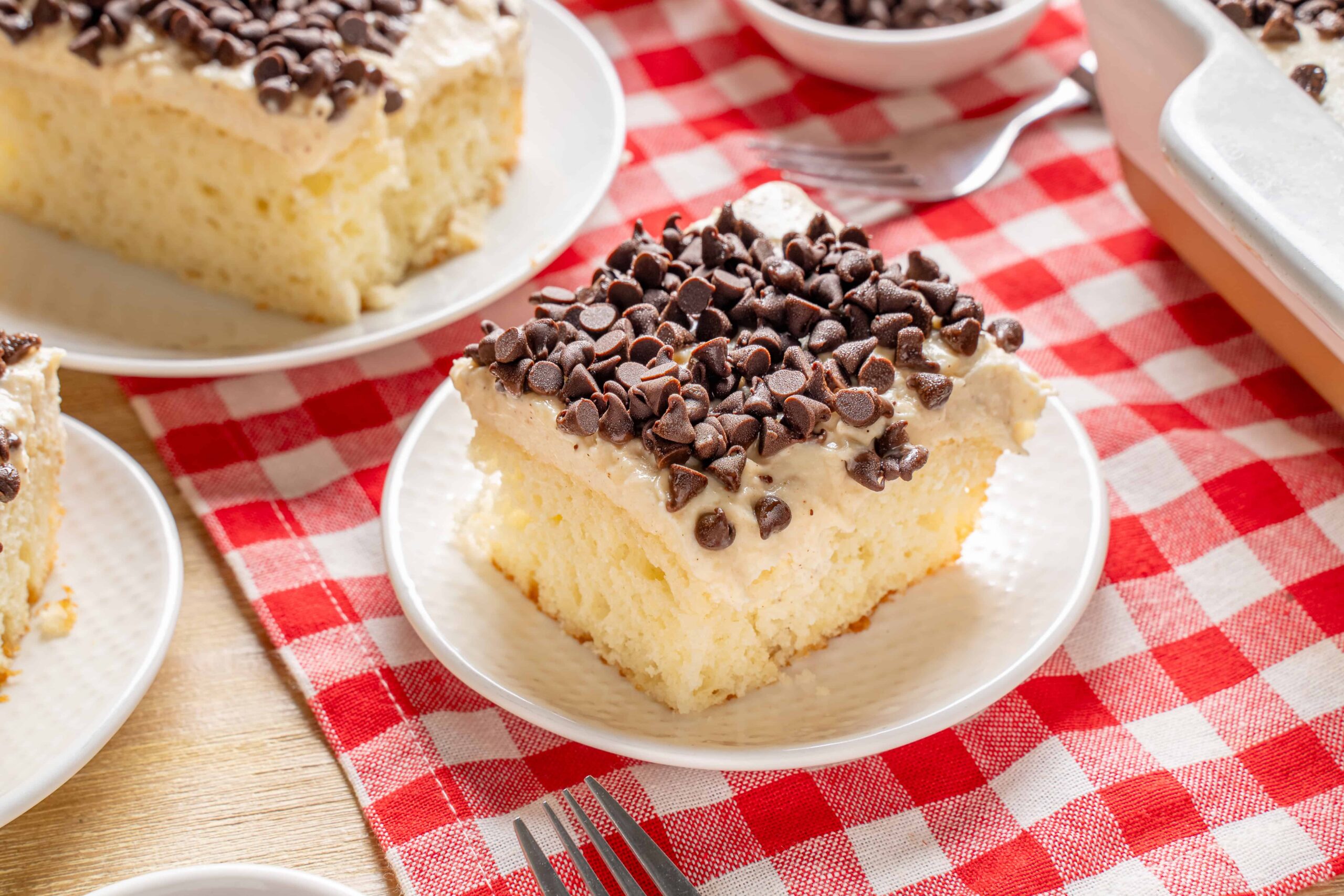 Creamy vanilla sponge cake topped with rich chocolate chips, served on a white plate with a red checkered tablecloth, perfect for dessert or teatime.