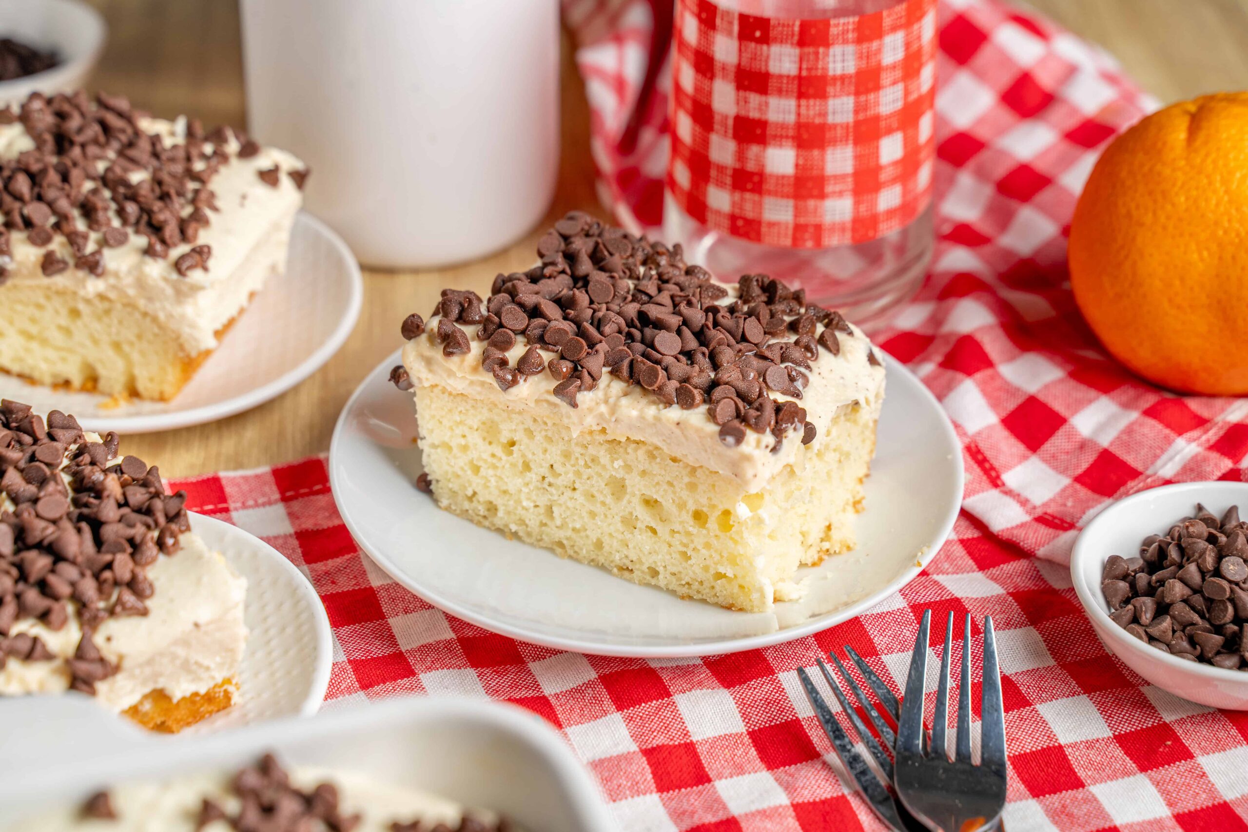 Creamy vanilla cake slice topped with rich chocolate chips on a white plate with a red checkered tablecloth, fresh orange, and dessert ingredients in the background.