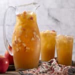 Fresh homemade apple juice in a glass pitcher and two glasses, with chopped apples inside, on a rustic wooden table against a light gray brick wall. Perfect for a healthy beverage or apple juice recipe.