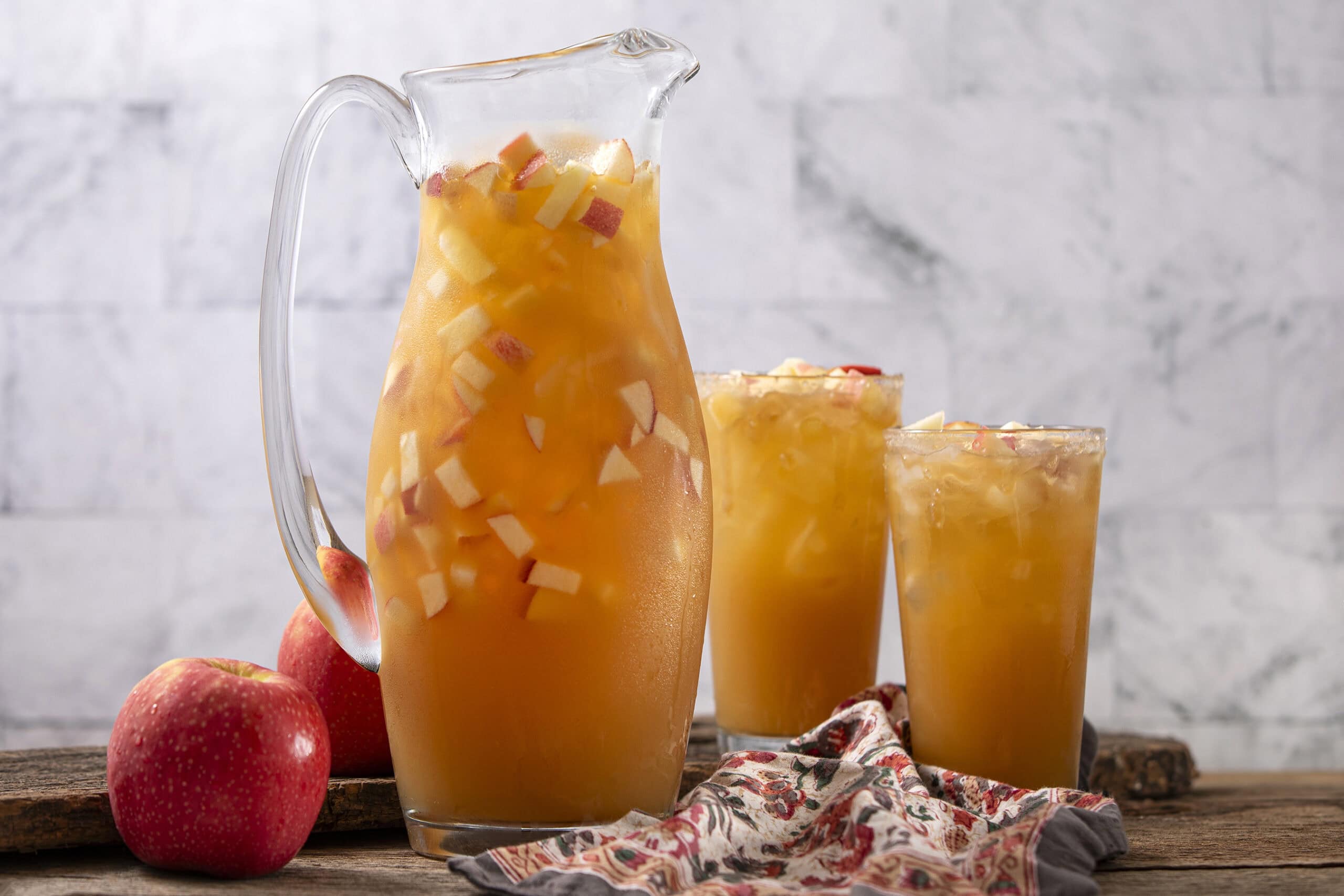 Fresh homemade apple juice in a glass pitcher and two glasses, with chopped apples inside, on a rustic wooden table against a light gray brick wall. Perfect for a healthy beverage or apple juice recipe.