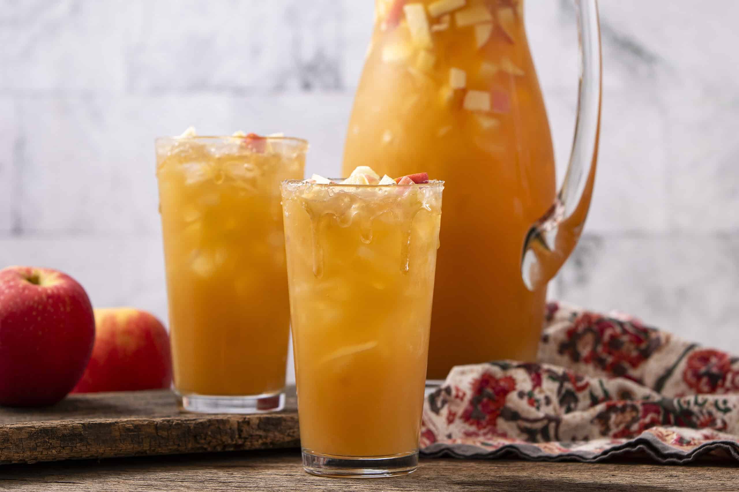 Fresh apple juice served in tall glasses with ice, with a pitcher of homemade apple cider in the background. Perfect for refreshing summer drinks and healthy beverage options.