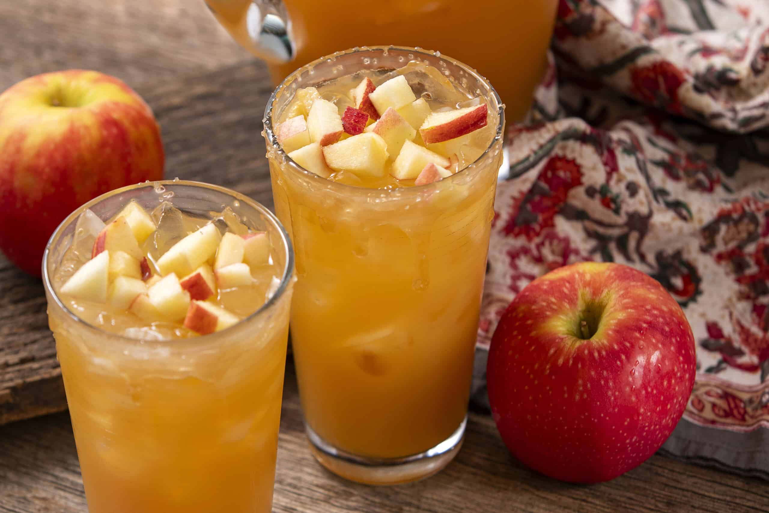 Fresh apple juice with chopped apples on top in tall glasses, surrounded by whole apples and a rustic wooden table for a healthy fruit drink presentation.