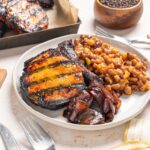 Succulent grilled ribeye steak with colorful vegetable toppings served with baked sweet potatoes and side dishes, showcasing a hearty, flavorful meal.