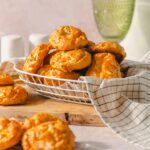 Fried cheese biscuits stacked in a basket with a checkered cloth, served on a wooden tray with a glass of water in the background, perfect for an appetizer or snack.