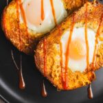 Golden-brown toast with baked eggs, drizzled with hot sauce on a black plate. A savory breakfast dish featuring crispy bread and perfectly cooked eggs, ideal for brunch or a hearty start to the day.