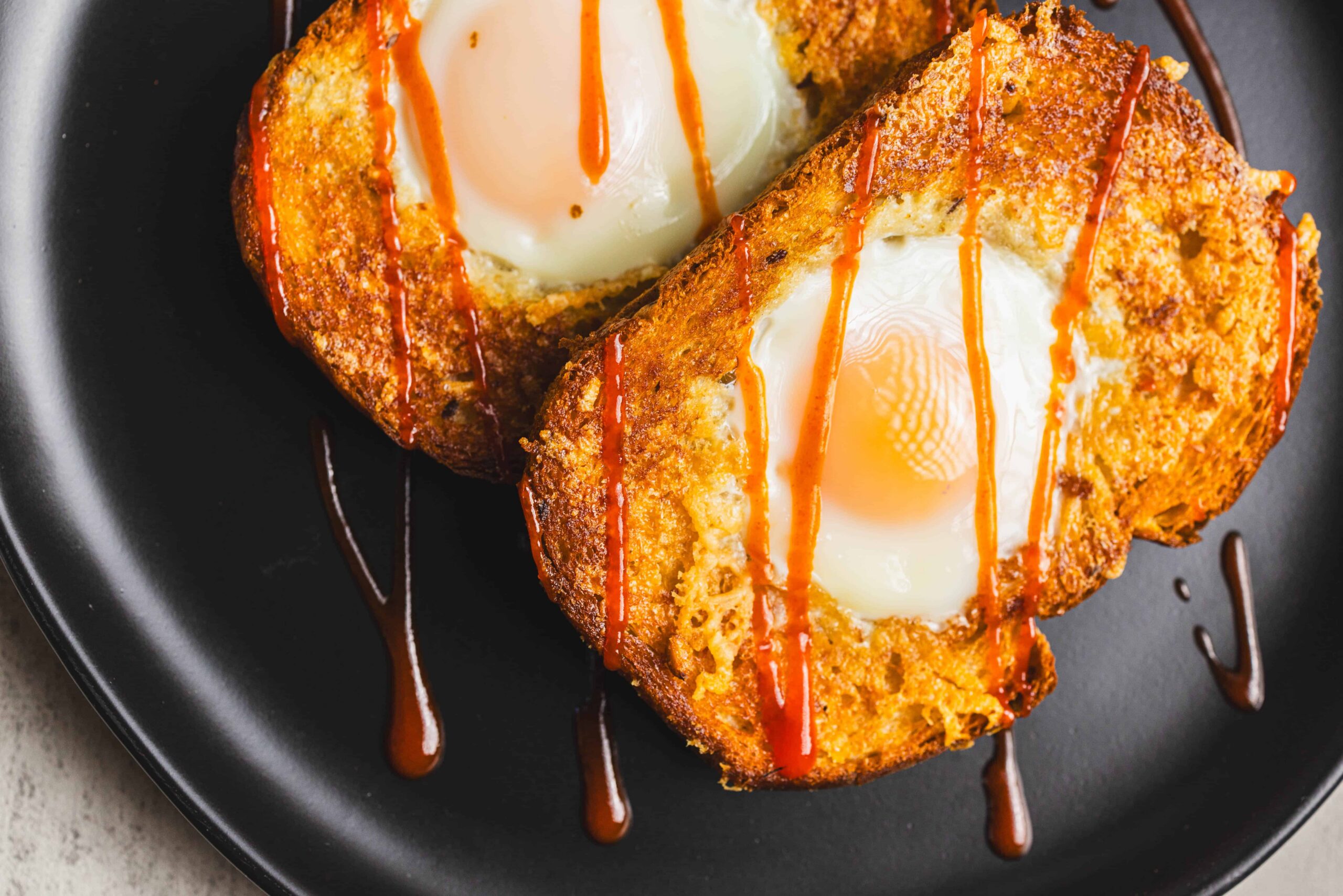 Golden-brown toast with baked eggs, drizzled with hot sauce on a black plate. A savory breakfast dish featuring crispy bread and perfectly cooked eggs, ideal for brunch or a hearty start to the day.