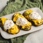 Baked sweet potatoes topped with poached eggs, melted cheese, and red pepper flakes, served on a white textured platter with a green cloth backdrop.