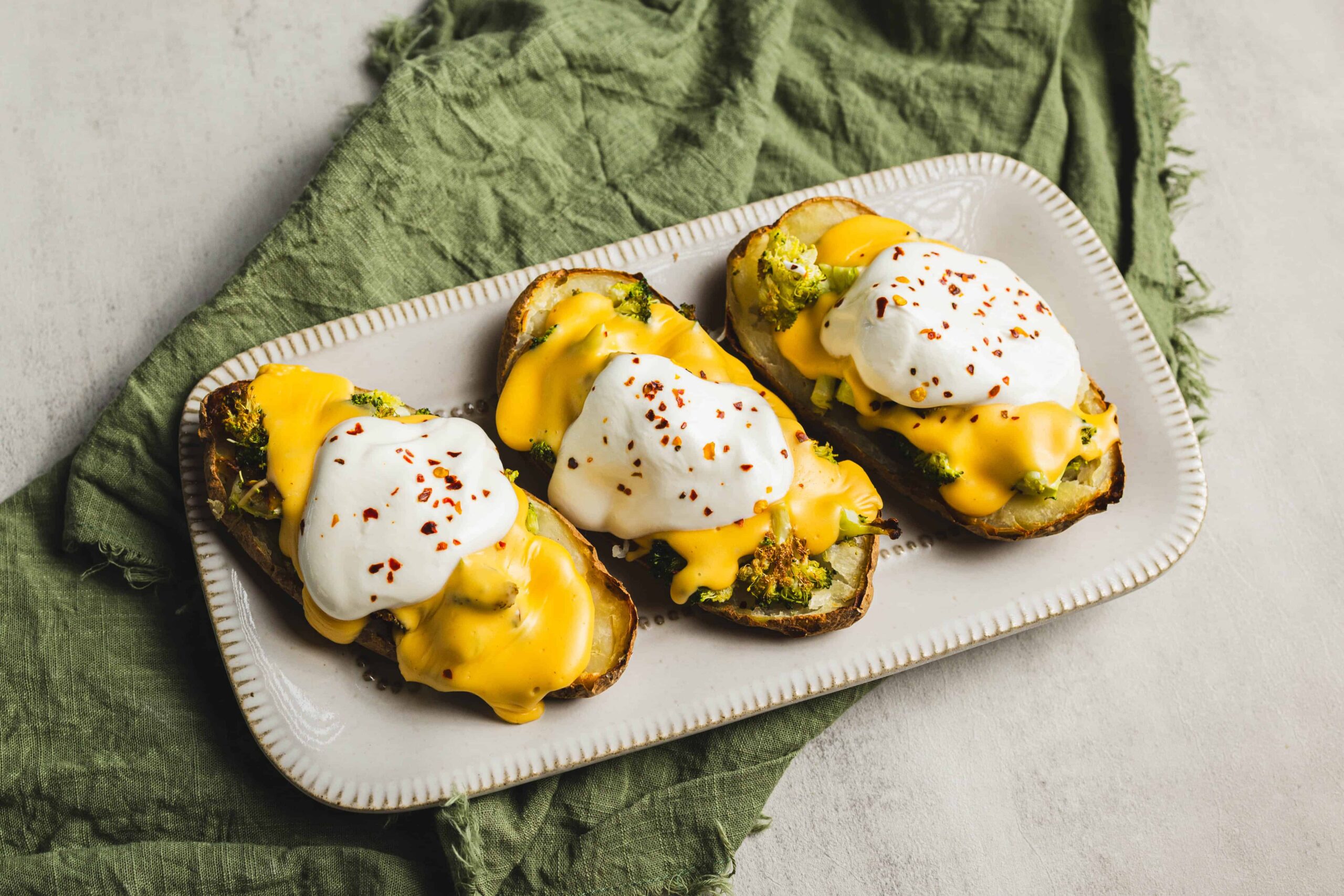 Baked sweet potatoes topped with poached eggs, melted cheese, and red pepper flakes, served on a white textured platter with a green cloth backdrop.