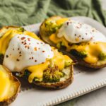 Roasted sweet potatoes topped with poached eggs, melted cheddar cheese, and sprinkled red pepper flakes on a white platter, served with a green cloth background.