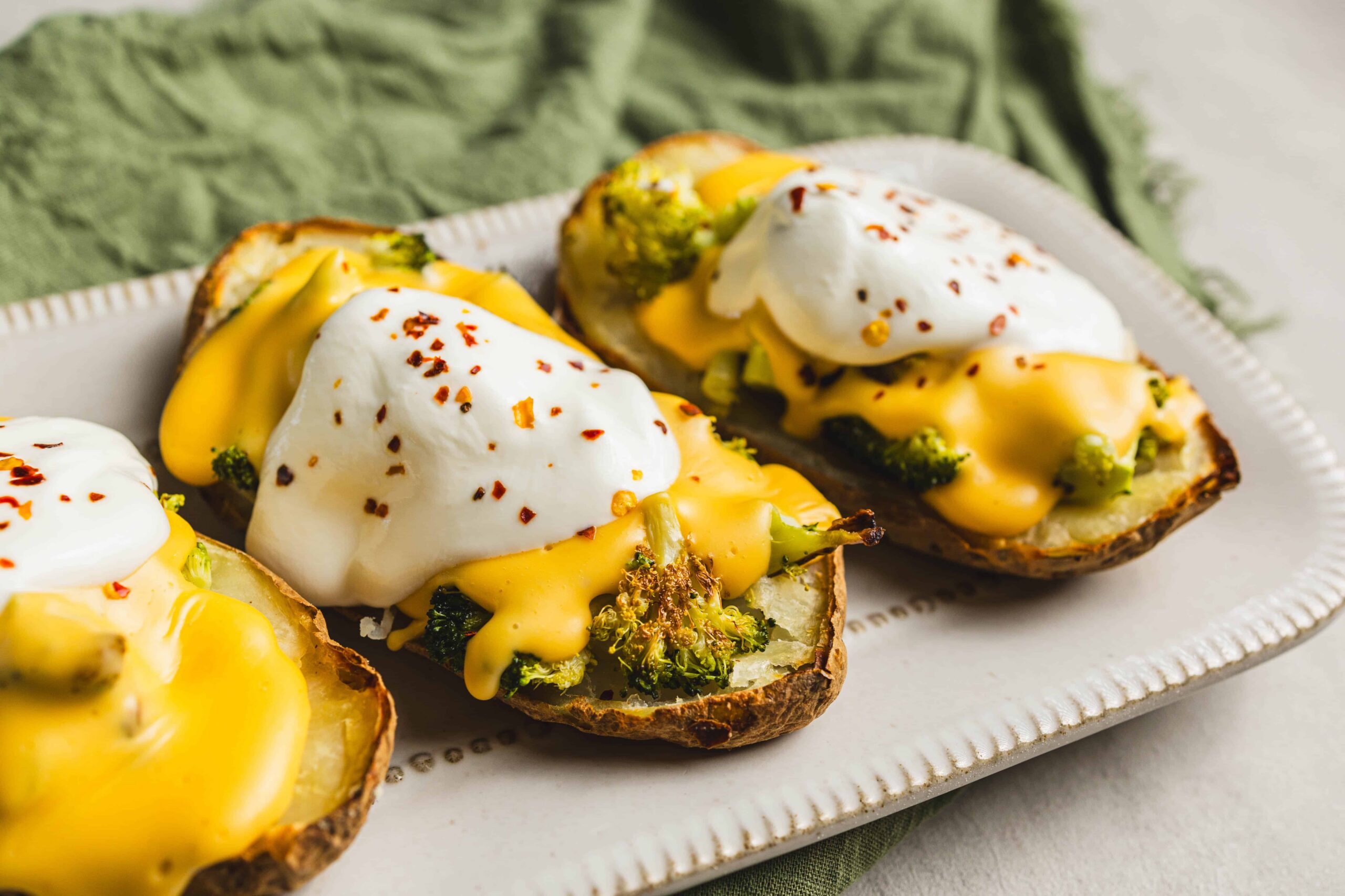 Roasted sweet potatoes topped with poached eggs, melted cheddar cheese, and sprinkled red pepper flakes on a white platter, served with a green cloth background.