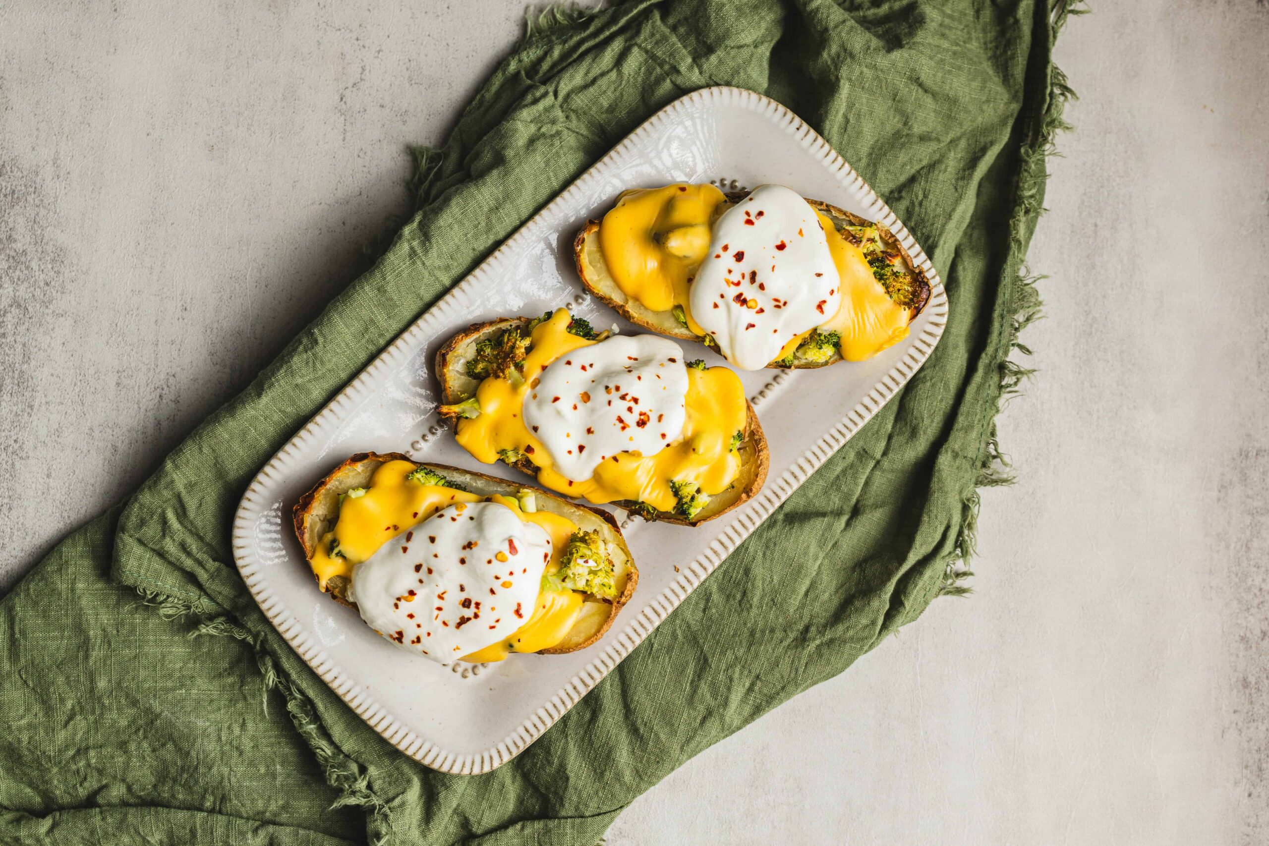 Savory breakfast eggs Benedict with poached eggs, hollandaise sauce, broccoli, and red pepper flakes on toasted bread slices served on a white platter with a green cloth background.