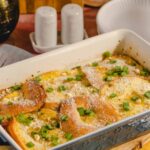 Golden baked breakfast casserole with cheese, green onions, and crispy bread slices, served in a ceramic baking dish with surroundings of salt and pepper shakers, plates, and utensils on a rustic wooden table.
