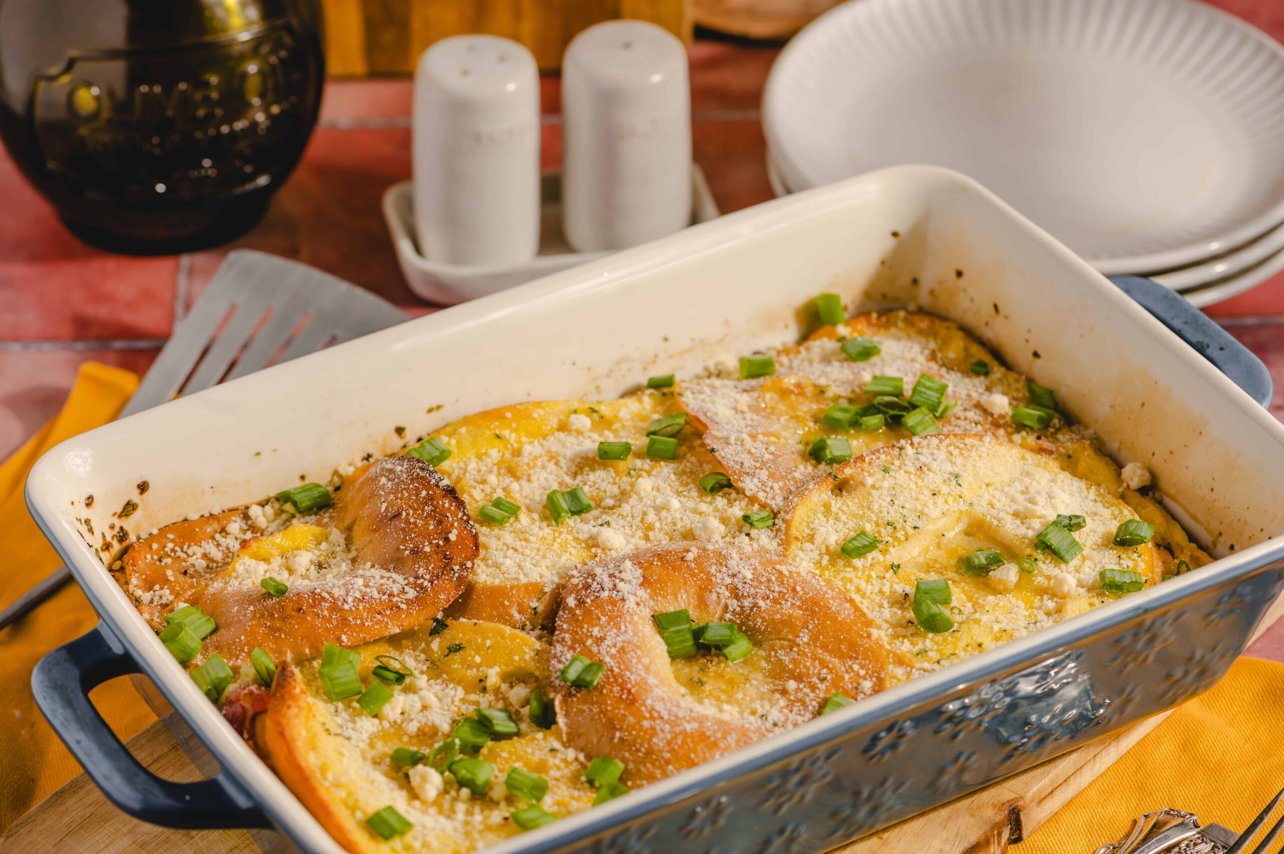 Golden baked breakfast casserole with cheese, green onions, and crispy bread slices, served in a ceramic baking dish with surroundings of salt and pepper shakers, plates, and utensils on a rustic wooden table.