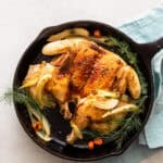 Roasted chicken with herbs and vegetables in a cast iron skillet, served with a glass of white wine, on a white surface with utensils and a light blue napkin.