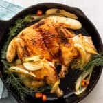 Golden roasted whole chicken in a skillet with caramelized onions, carrots, and fresh herbs, served with a glass of white wine for a delicious and flavorful meal.