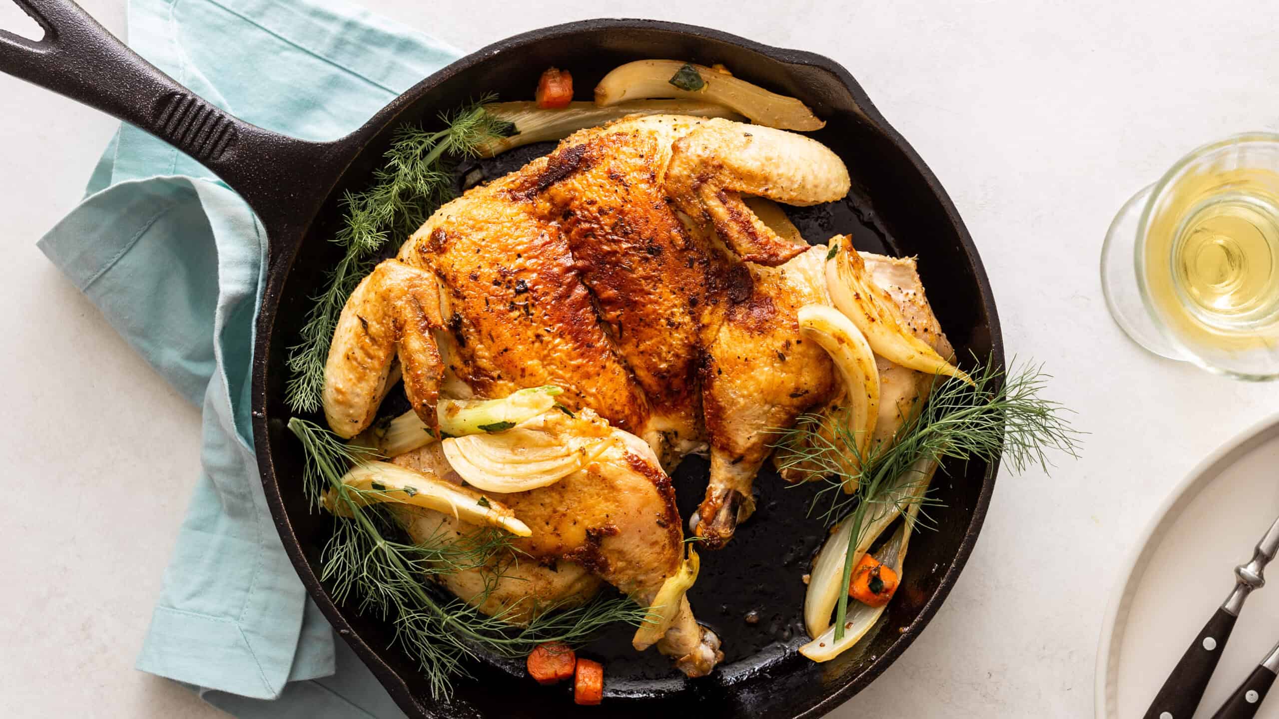 Golden roasted whole chicken in a skillet with caramelized onions, carrots, and fresh herbs, served with a glass of white wine for a delicious and flavorful meal.