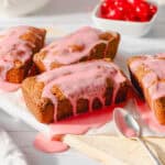 Freshly baked pound cakes drizzled with pink cherry glaze, served on a white wooden surface, with cherries in a bowl in the background, perfect for dessert recipes and cherry-flavored treats.