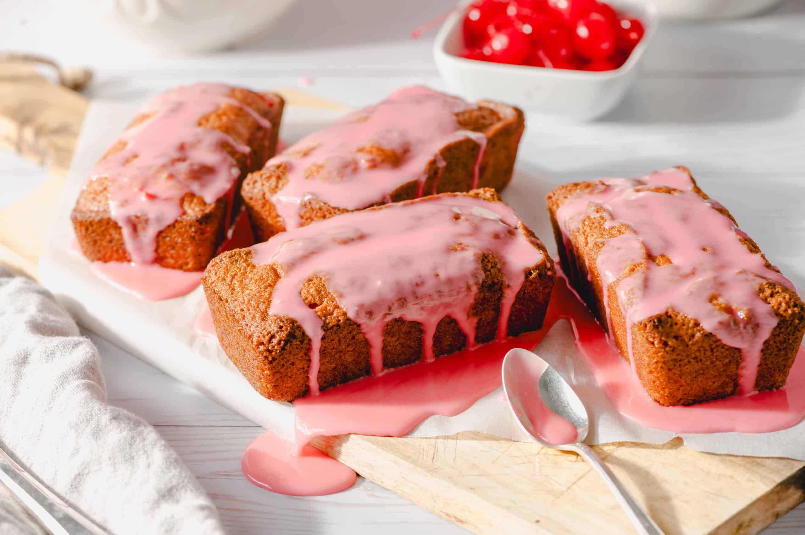 Freshly baked pound cakes drizzled with pink cherry glaze, served on a white wooden surface, with cherries in a bowl in the background, perfect for dessert recipes and cherry-flavored treats.