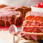 Moist strawberry cake with pink glaze drizzled on top, sliced to reveal soft, fluffy interior, served on parchment paper with fresh strawberry topping in the background.