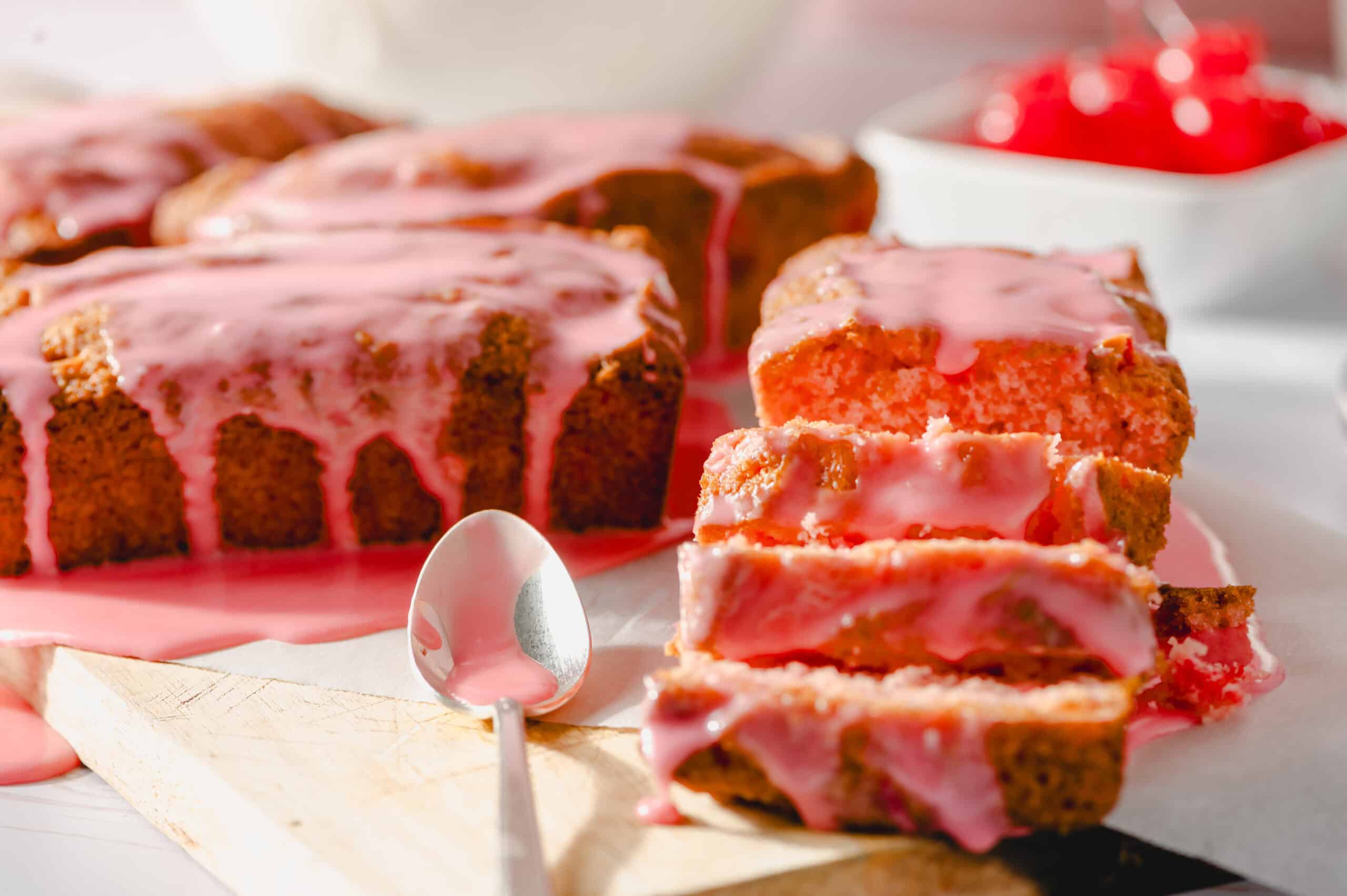 Moist strawberry cake with pink glaze drizzled on top, sliced to reveal soft, fluffy interior, served on parchment paper with fresh strawberry topping in the background.