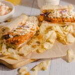 Tender fried chicken sandwich with coleslaw on a sub roll, served with crispy potato chips on a wooden cutting board, perfect for a delicious meal.