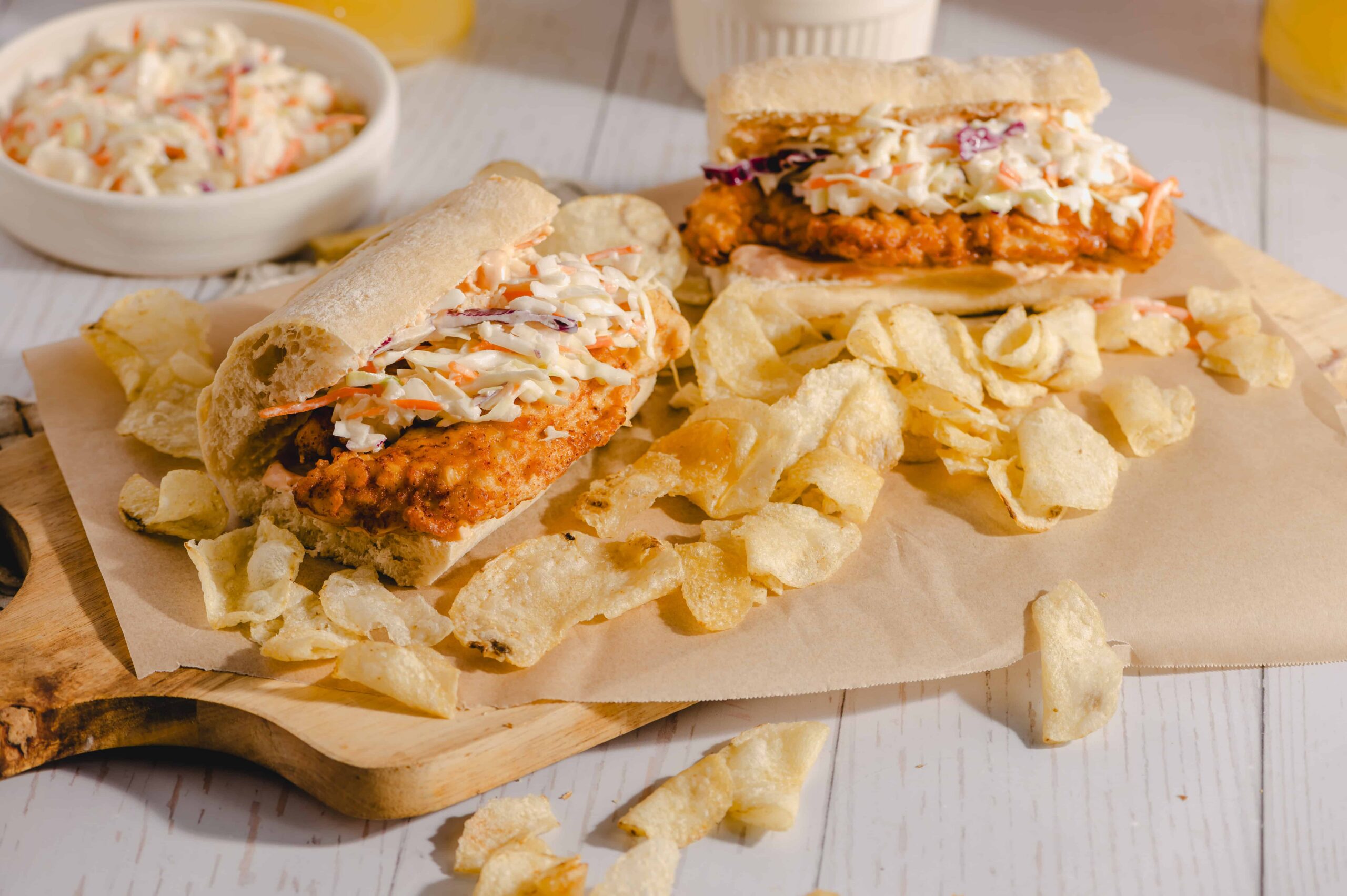 Tender fried chicken sandwich with coleslaw on a sub roll, served with crispy potato chips on a wooden cutting board, perfect for a delicious meal.