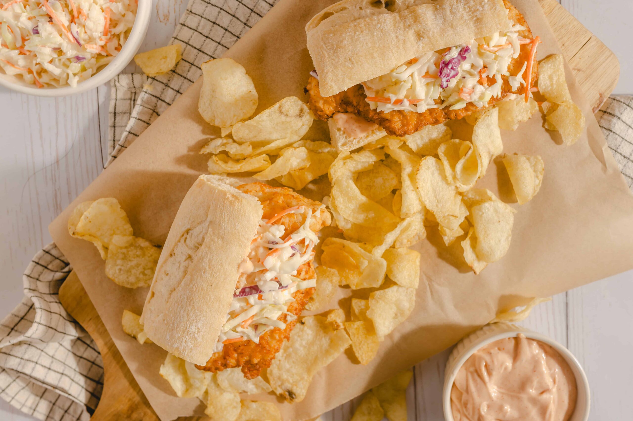 Decadent fried chicken sandwich with coleslaw and potato chips, served with creamy dipping sauce on a rustic wooden board. Perfect comfort food featuring crispy chicken, fresh coleslaw, and crunchy chips.
