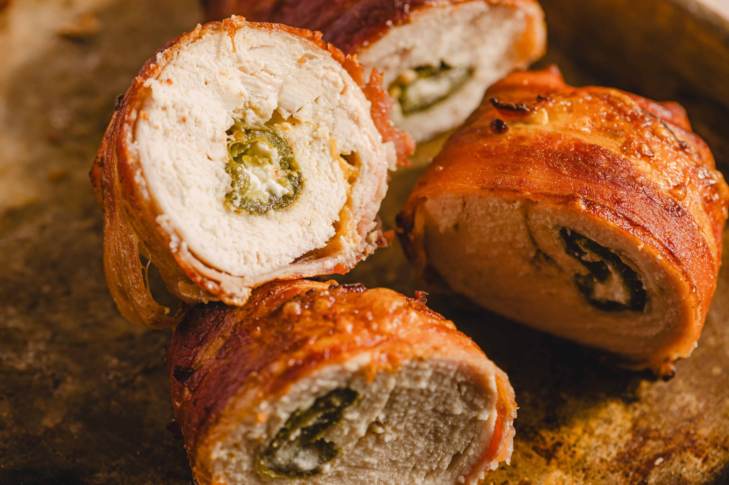 Juicy chicken breast roulades stuffed with cheese and jalapeno peppers, wrapped in crispy bacon, cooked to perfection, and sliced to reveal flavorful filling, served on a rustic baking sheet.