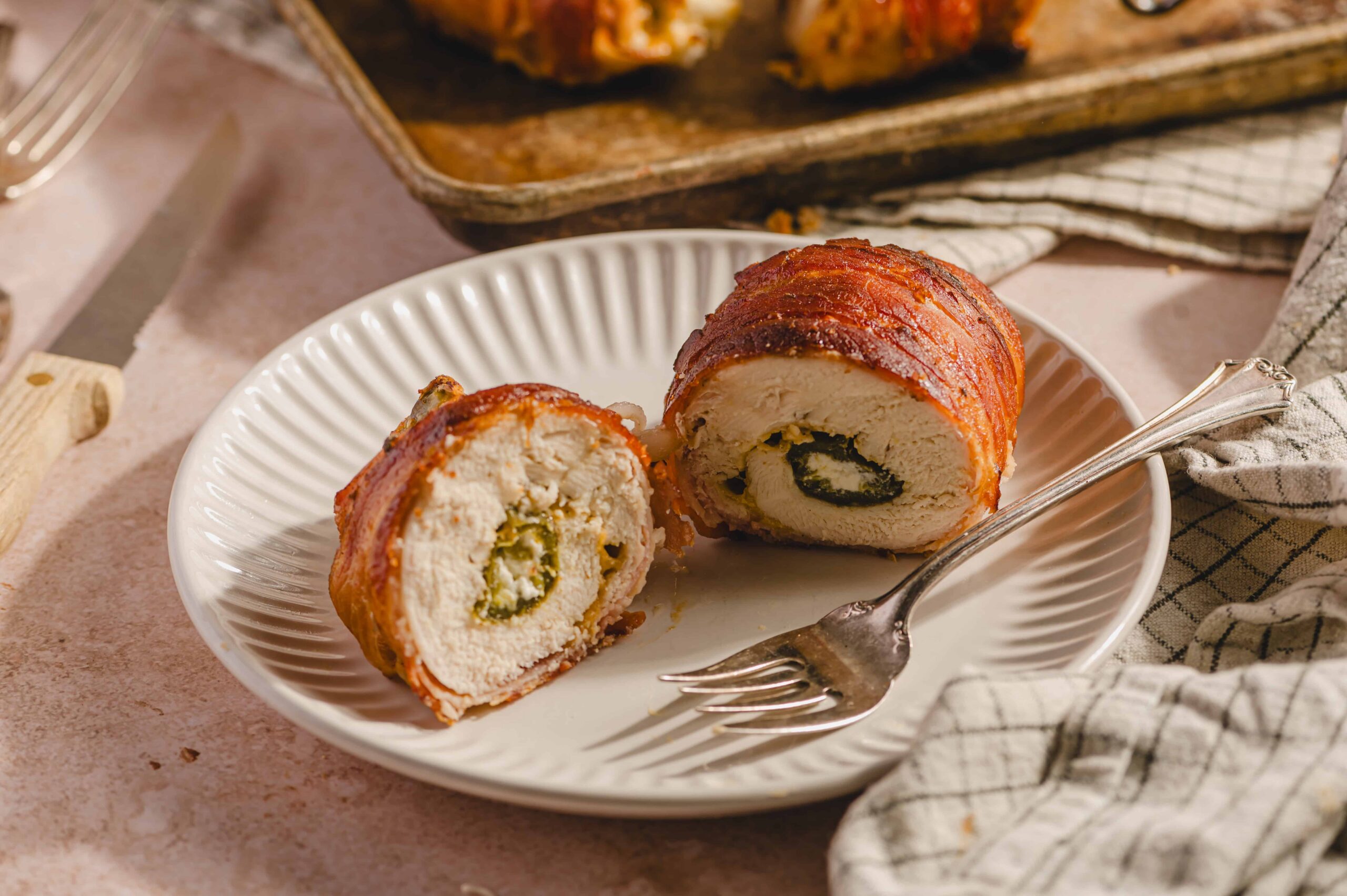 Tender chicken cordon bleu stuffed with cheese and ham, wrapped in crispy bacon, sliced open to reveal a savory filling, served on a elegant white plate with a vintage fork on a rustic kitchen table.