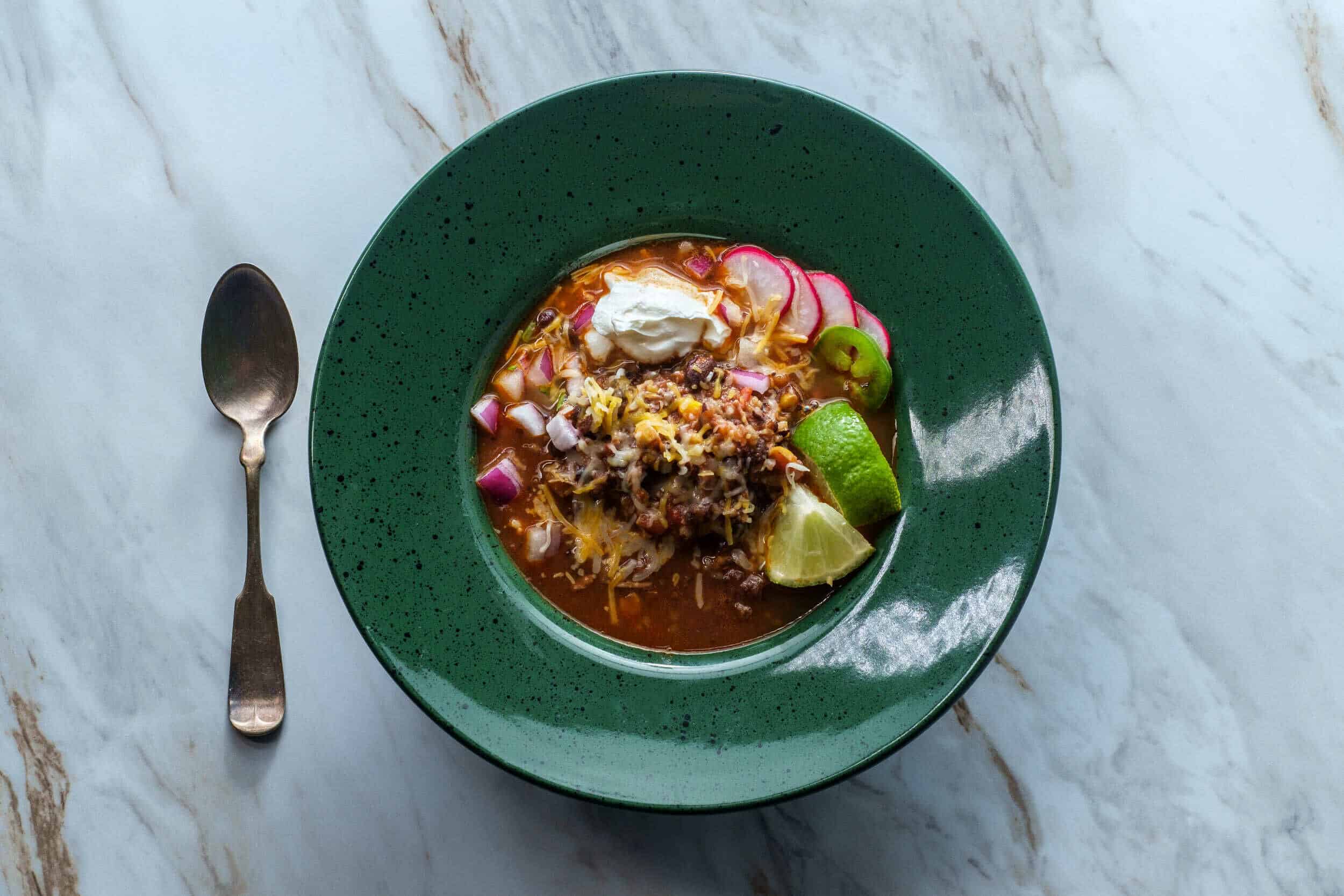 Savory bowl of Mexican pozole with shredded meat, radishes, lime wedges, shredded cheese, sour cream, and chopped onions served in a green bowl.