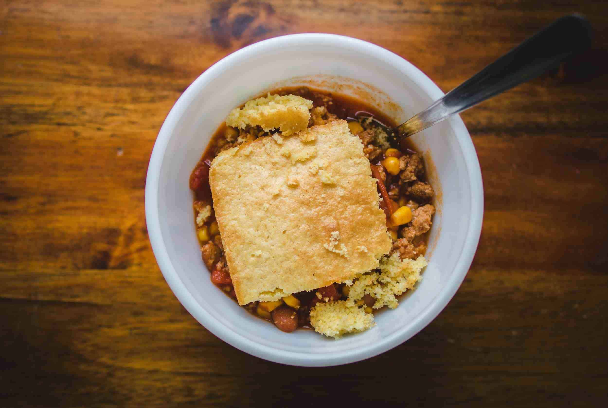 Creamy beef chili with corn and shredded cheese in a white bowl on a wooden table, topped with a piece of cornbread.