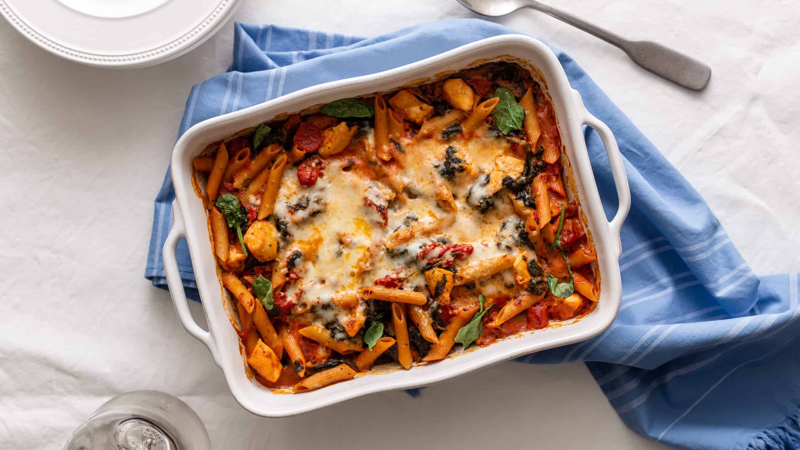 Creamy baked pasta casserole with melted cheese, herbs, and colorful vegetables in a white baking dish on a light table. Perfect comfort food recipe for family dinners and meal prep.