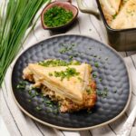 Savory chicken pot pie with flaky pastry crust garnished with chopped chives on a textured black plate. Includes fresh herbs, sliced green onions, and a casserole dish with cooked chicken pot pie in the background.