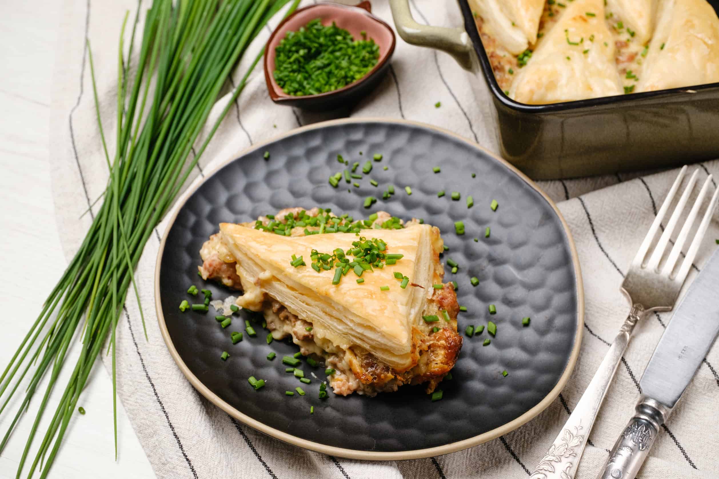 Savory chicken pot pie with flaky pastry crust garnished with chopped chives on a textured black plate. Includes fresh herbs, sliced green onions, and a casserole dish with cooked chicken pot pie in the background.