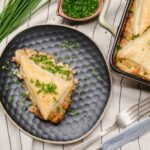 Creamy chicken pie with a golden flaky crust garnished with chopped chives on a black textured plate and baking dish, served with fresh chives on a striped cloth.