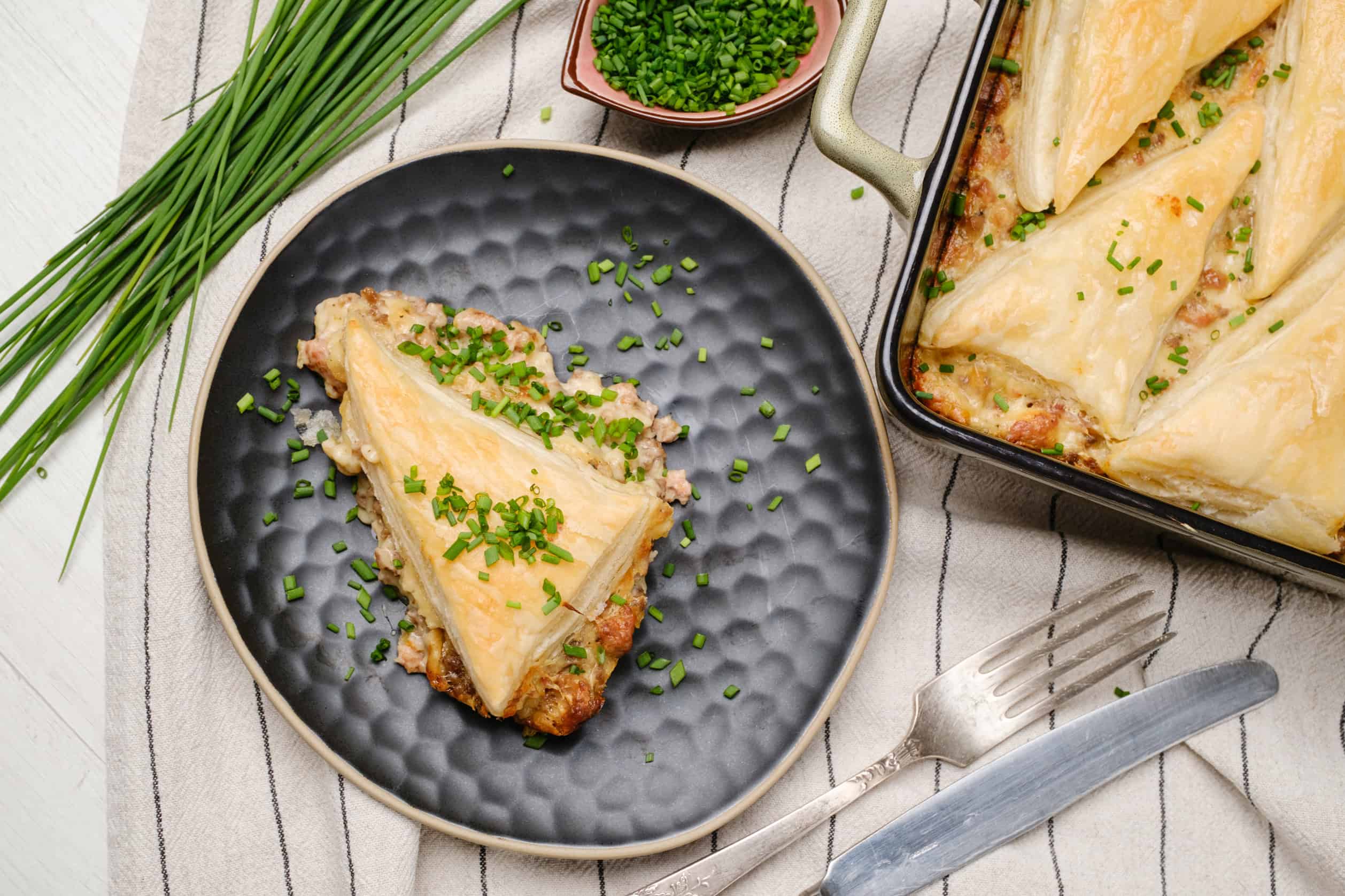 Creamy chicken pie with a golden flaky crust garnished with chopped chives on a black textured plate and baking dish, served with fresh chives on a striped cloth.