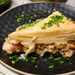 Creamy chicken pot pie with flaky pastry crust on black ceramic plate, garnished with chopped green onions, served with fresh herbs on side, comfort food idea.