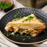 Creamy chicken and mushroom pot pie with a flaky puff pastry crust garnished with chopped chives, served on a stylish black plate. Perfect comfort food with savory filling and golden crust.